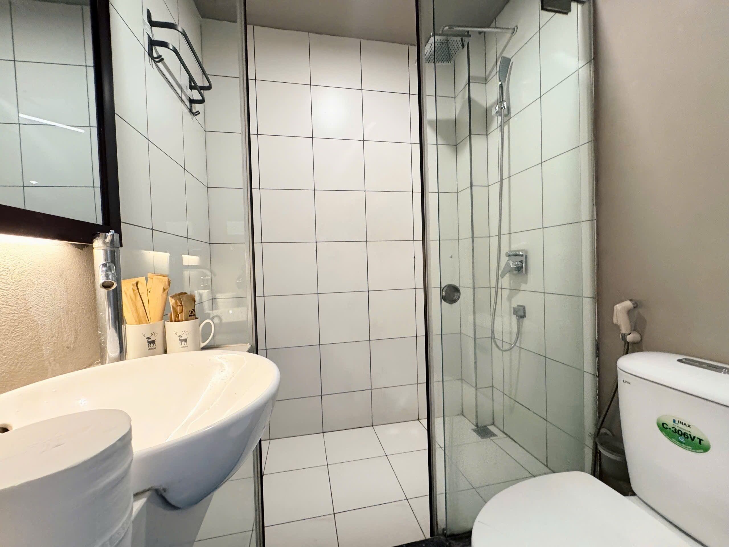 Un baño con lavamanos blanco, cabina de ducha con vidrio y un inodoro con un adhesivo verde.