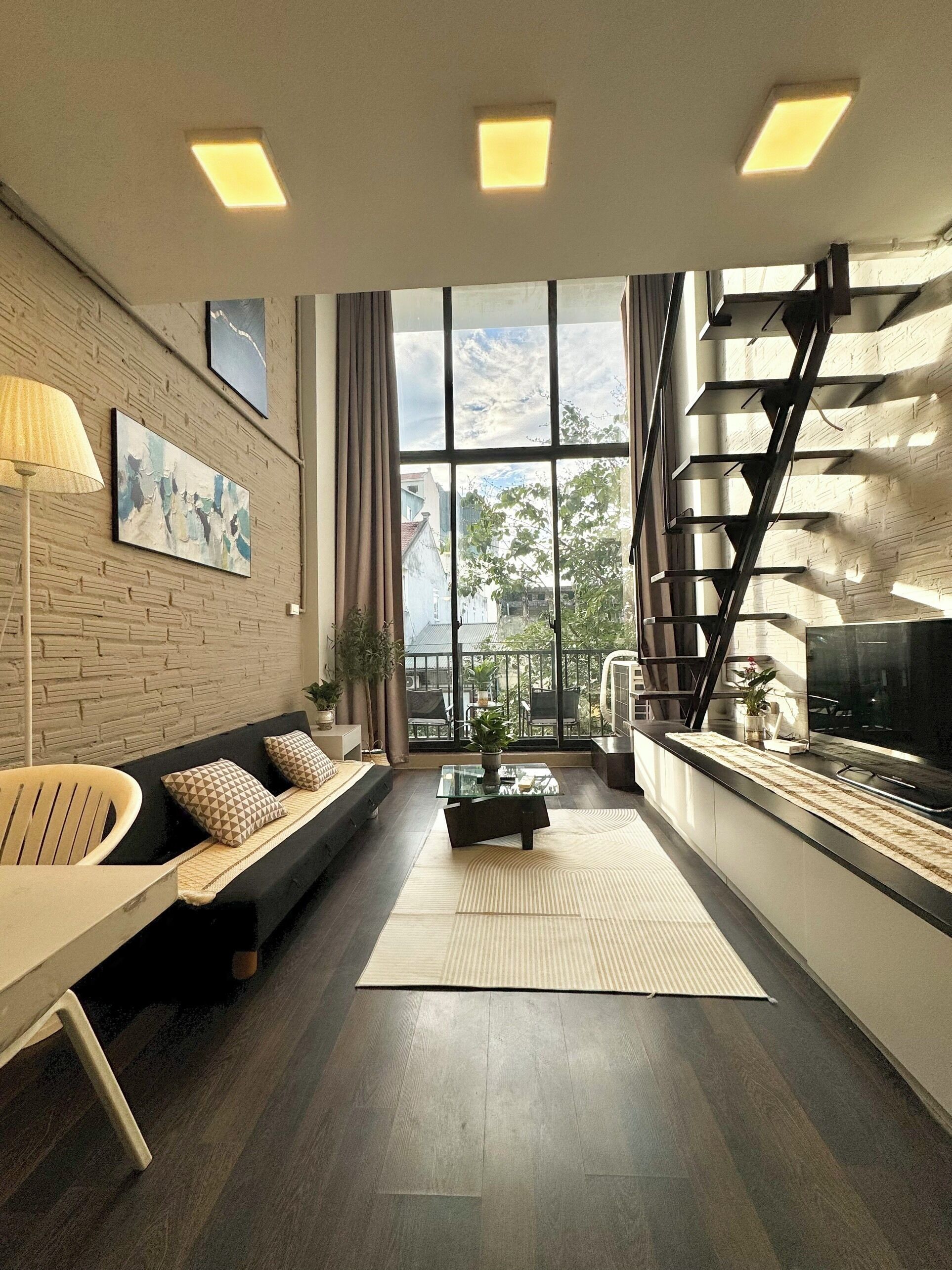 A modern living room with a sofa, coffee table, and a TV on a long counter. There are large windows with a view of the outside.