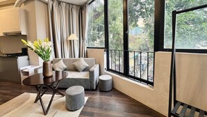 Una sala de estar moderna con un sofá, un reposapiés y una mesa con un jarrón de flores. Un ventanal grande ofrece vistas a la vegetación exterior.