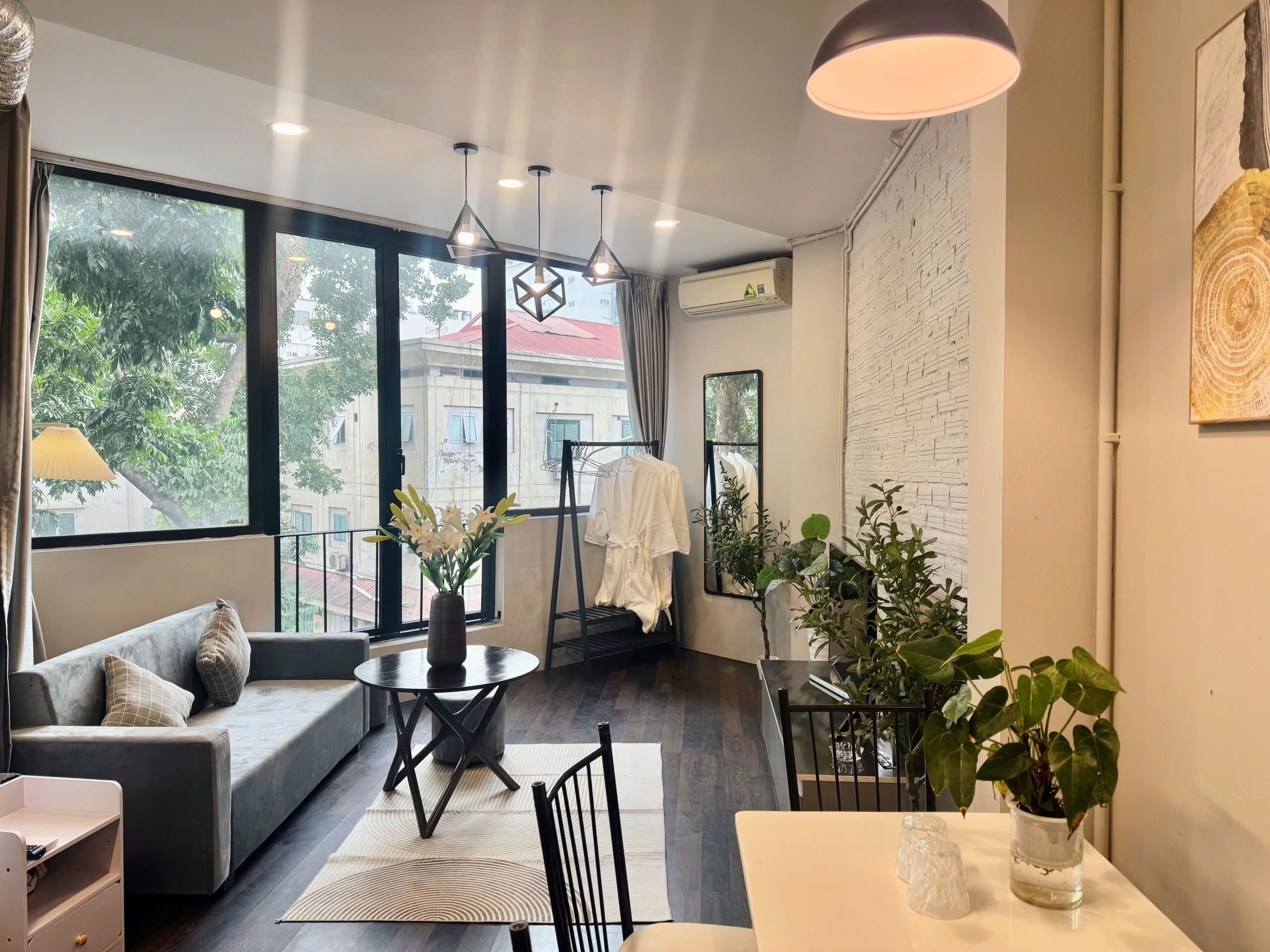A modern living room with a sofa, a round table, and a dining table. There are plants, a hanging light fixture, and a view of the outside through large windows.