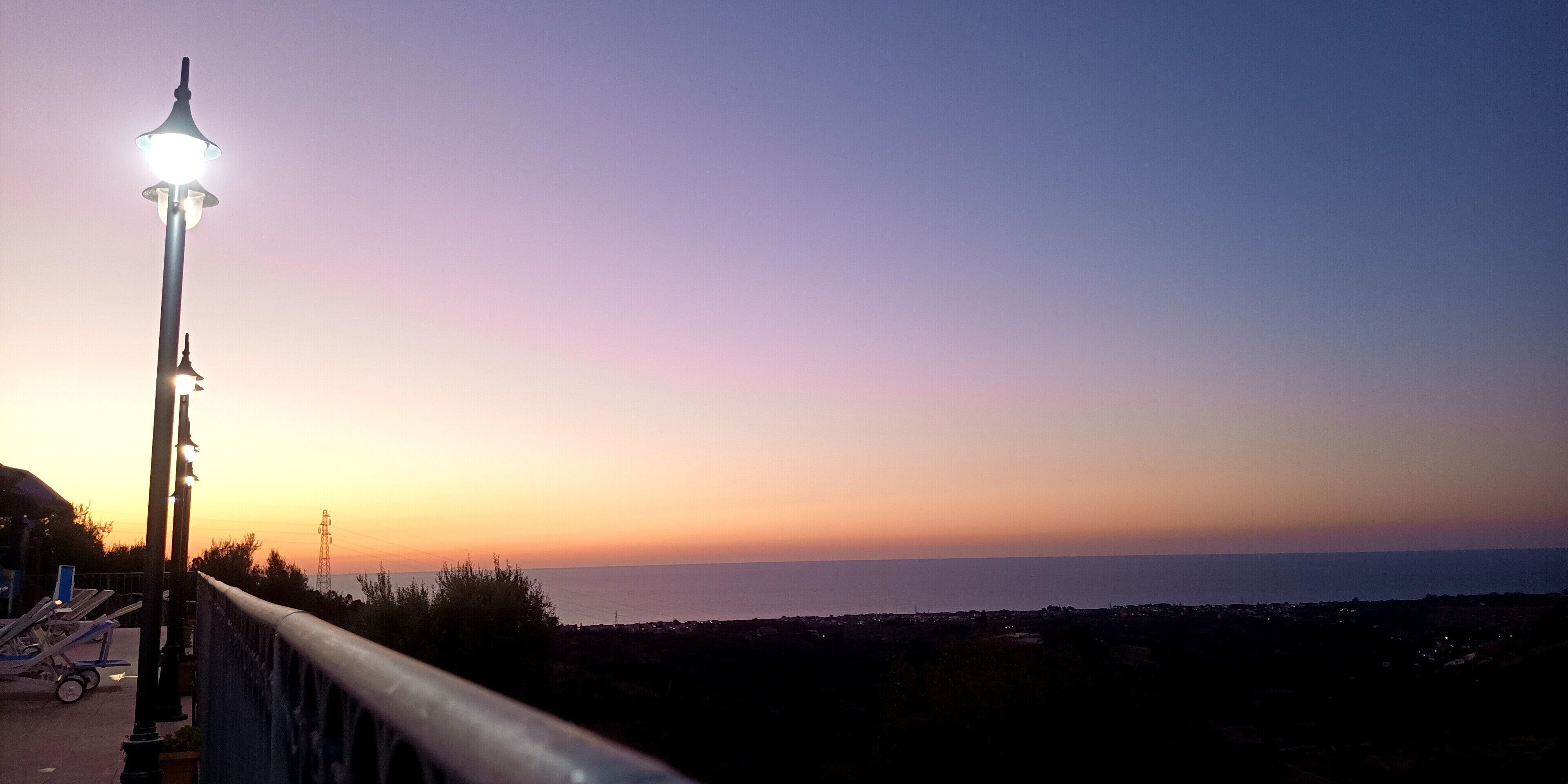 A sunset view from a balcony with a railing, overlooking a body of water and a distant shoreline.