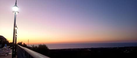 A sunset view from a balcony with a railing, overlooking a body of water and a distant shoreline.