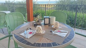 Une table ronde recouverte d’un plateau en mosaïque, entourée de chaises vertes, dressée pour un repas avec vue sur la mer et un paysage verdoyant en arrière-plan.