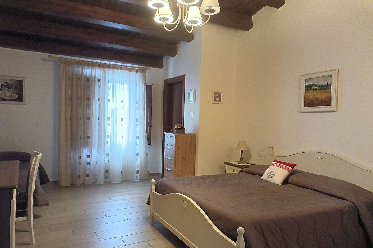 Un dormitorio con cama, cabecera de madera, mesita de noche, lámpara, silla, escritorio y ventana con cortinas.