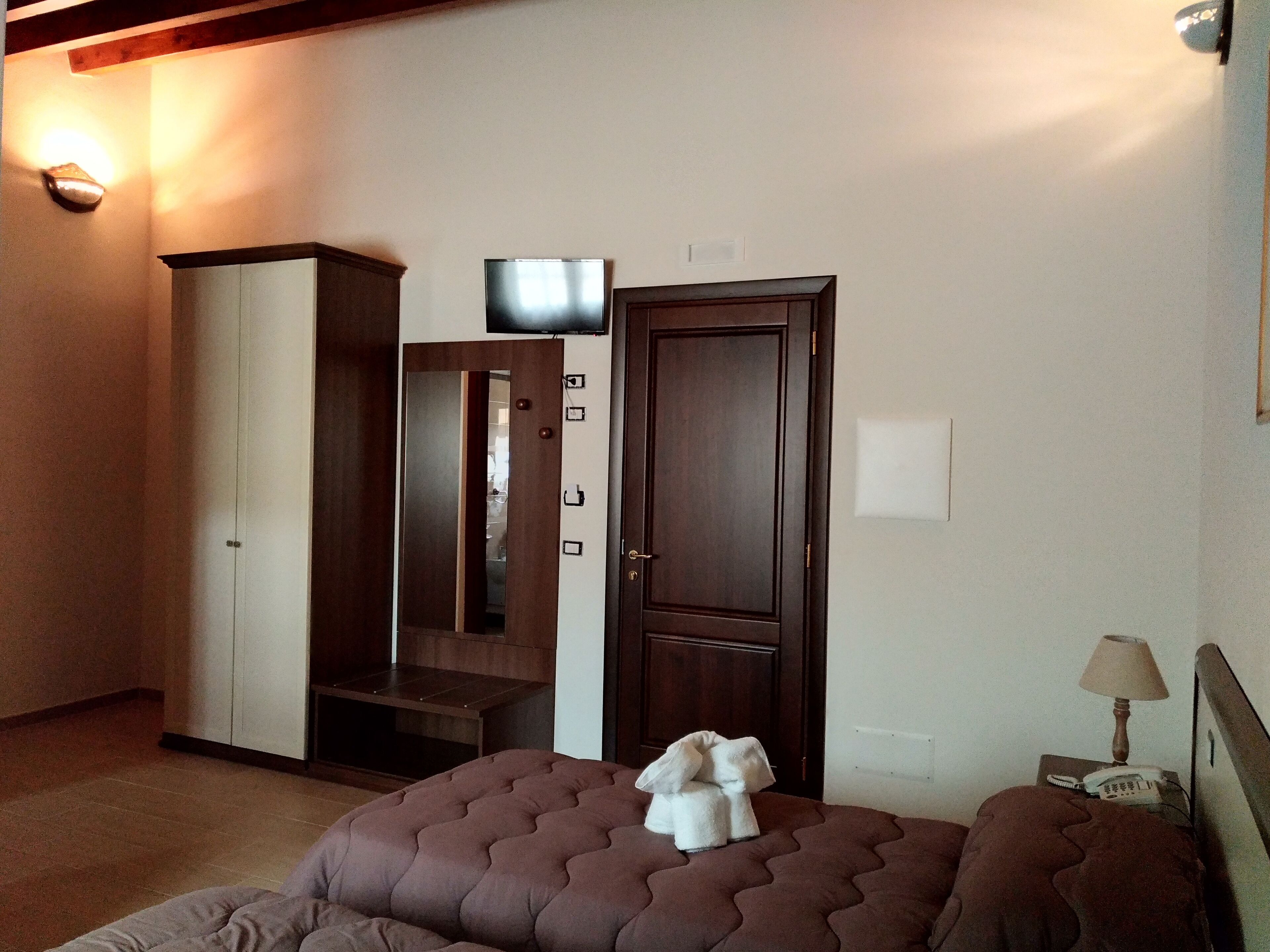 A hotel room with a bed, a wardrobe, a television, and a bedside table with a lamp and phone.