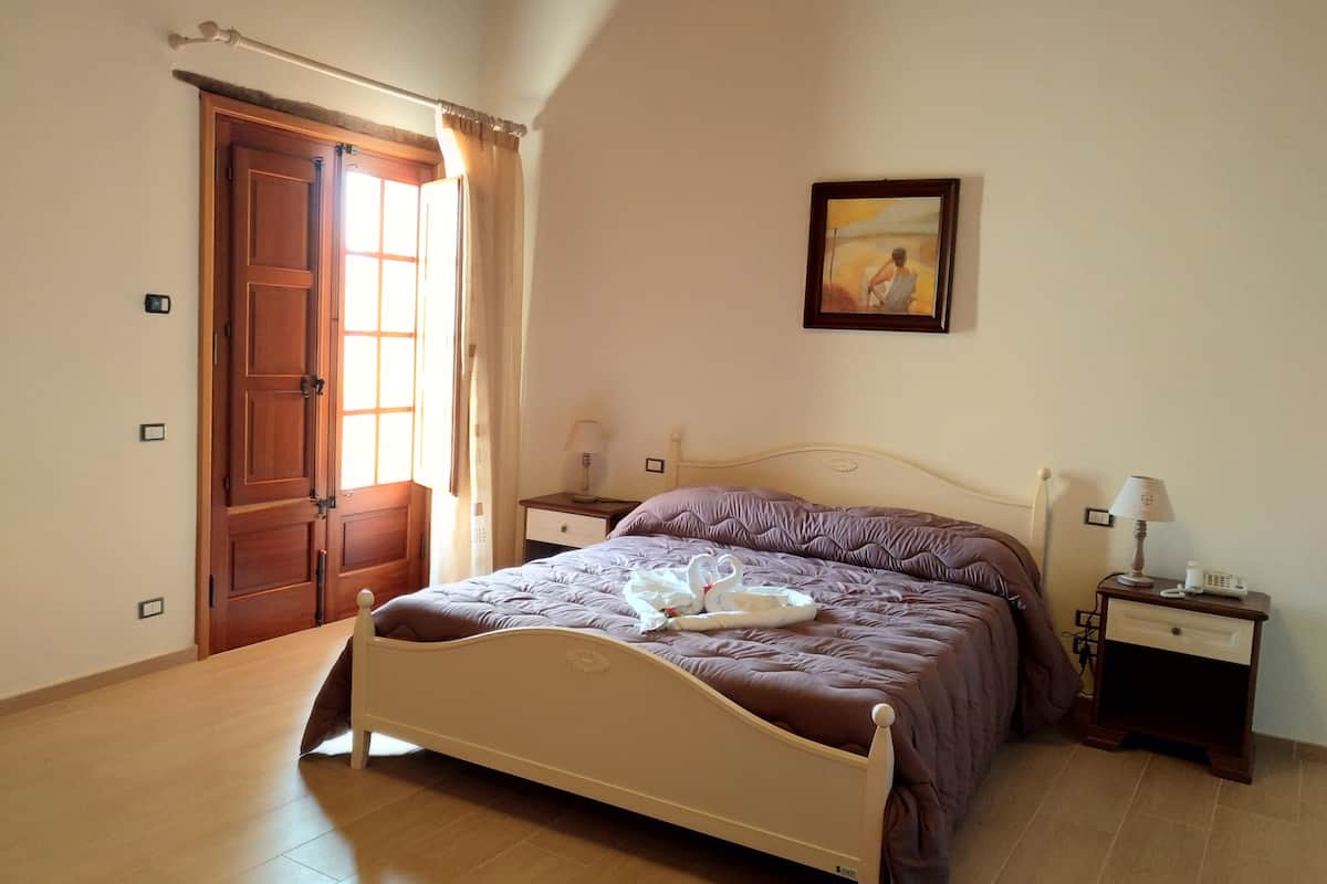 Un dormitorio con una cama de madera, una mesita de noche con lámpara, un cuadro en la pared y una puerta con cortinas.