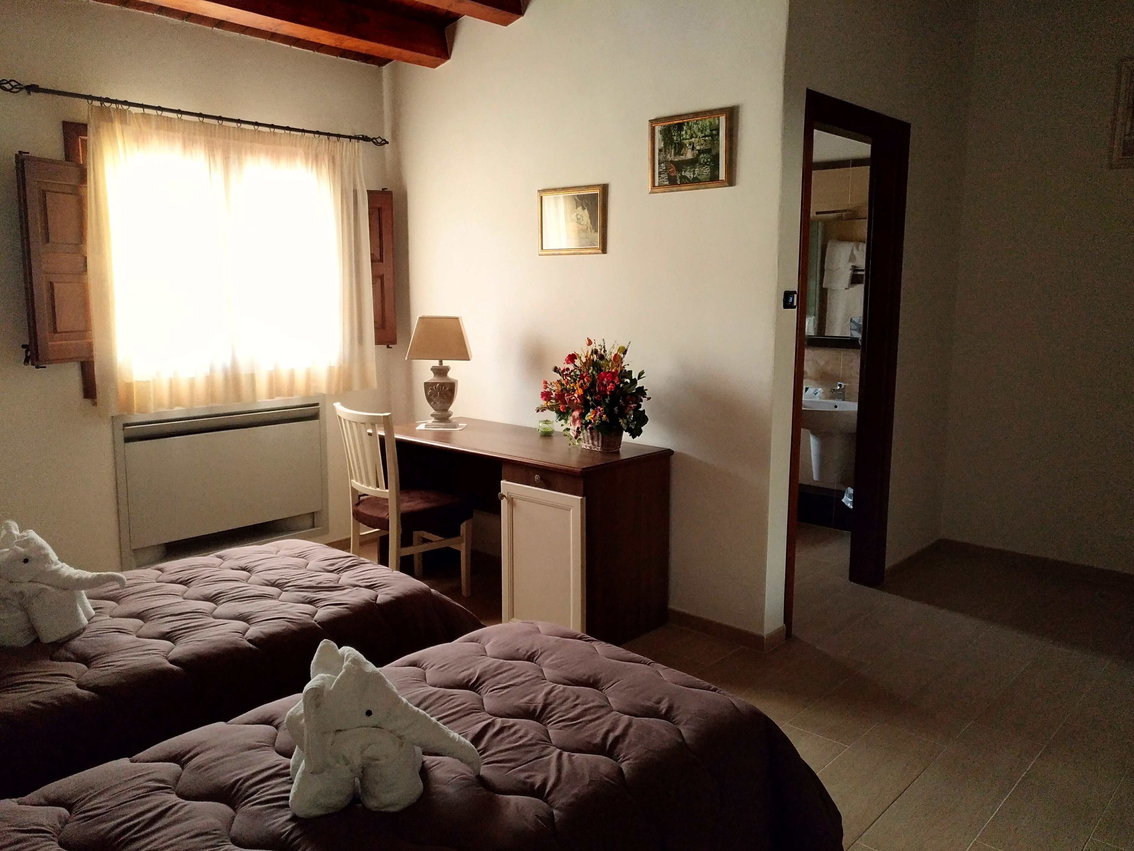 Habitación de hotel con dos camas, un escritorio con lámpara, una silla, un archivador pequeño y un jarrón con flores.