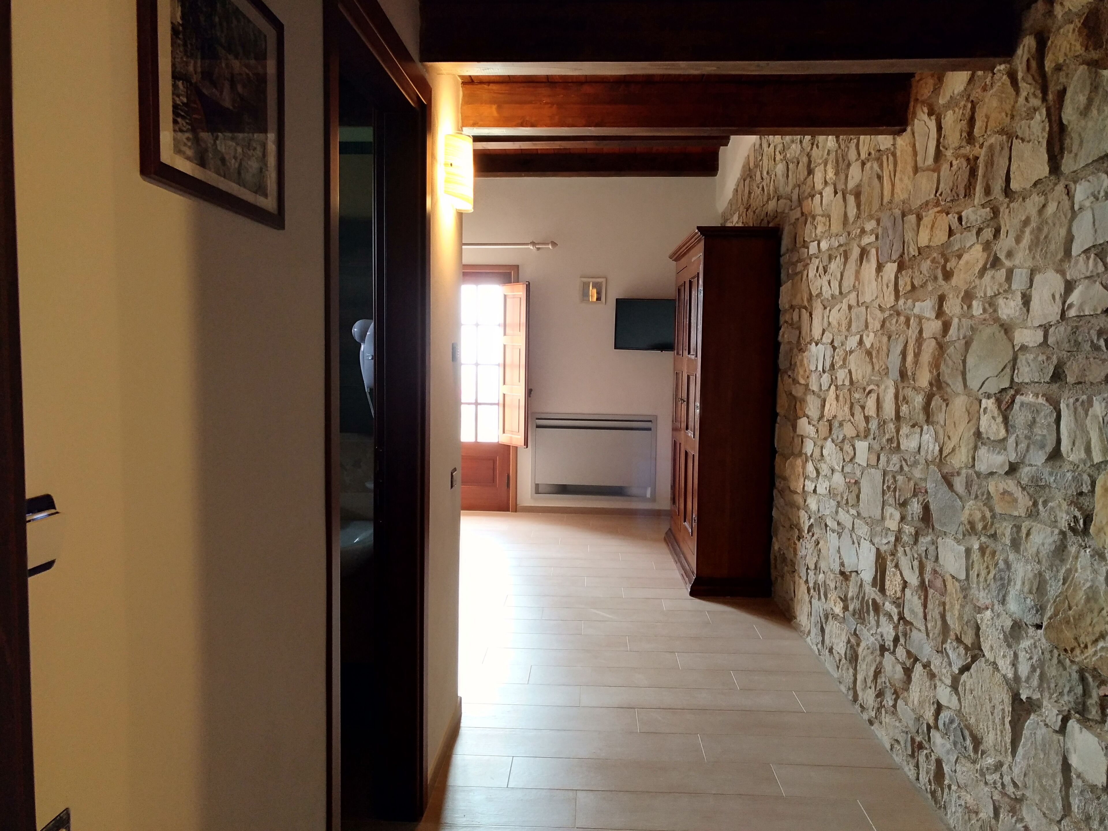 A hallway with a stone wall, wooden beams, and a tiled floor leading to a room with a window and a flat-screen TV.