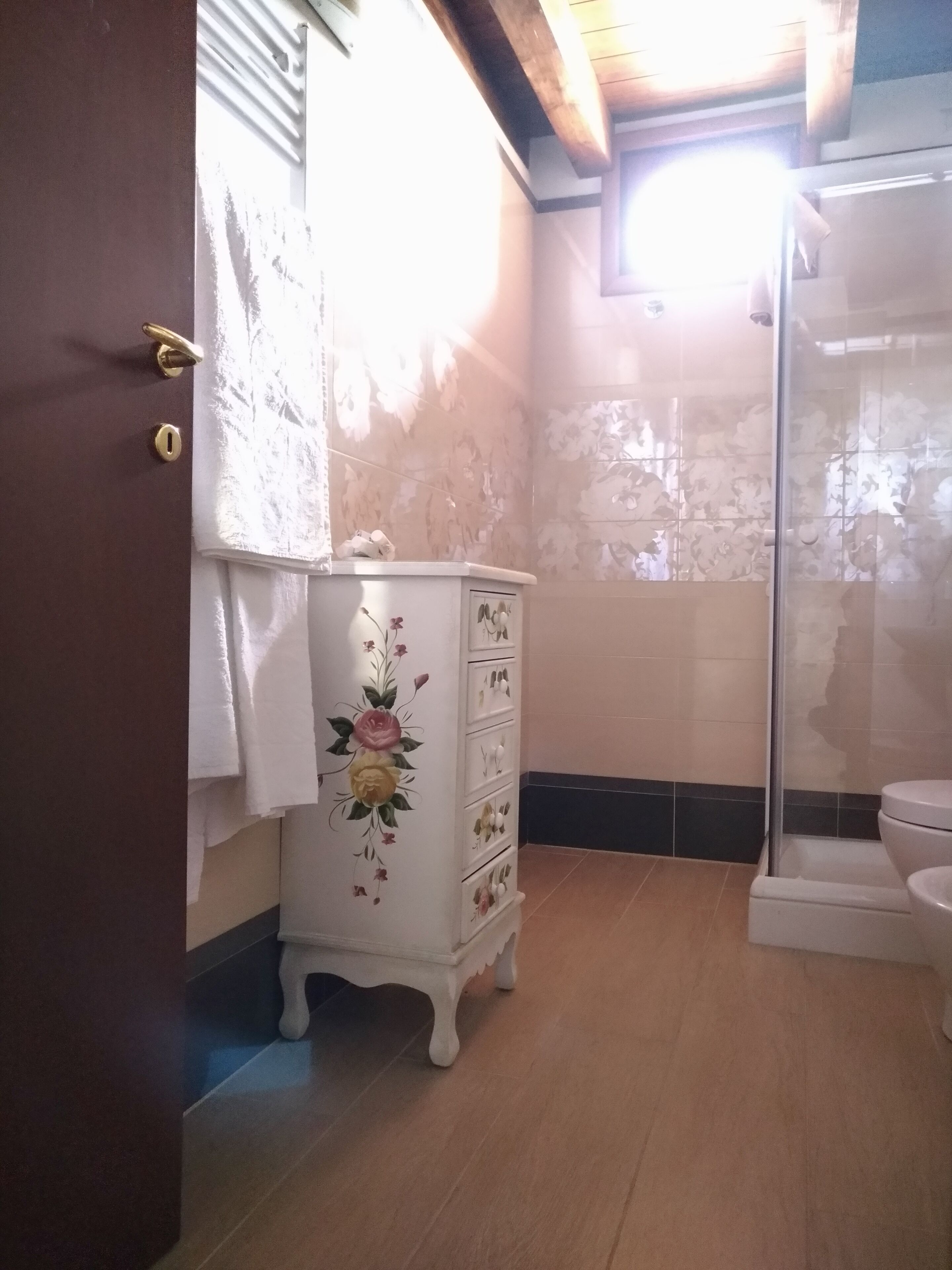 Un baño con piso de madera, un mueble blanco con diseños florales, un inodoro y una zona de ducha.