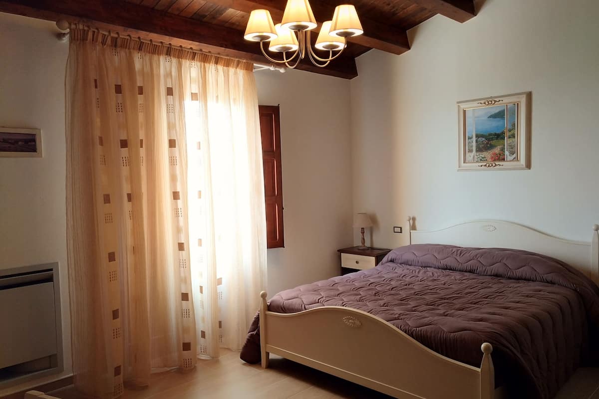 Un dormitorio con cama, mesita de noche, lámpara, un cuadro en la pared y ventana con cortinas.