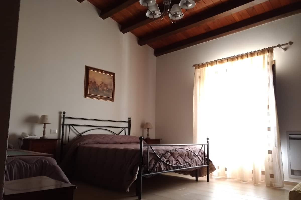 Un dormitorio con cama, una silla, una mesita de noche, una lámpara, un cuadro y una ventana con cortinas.