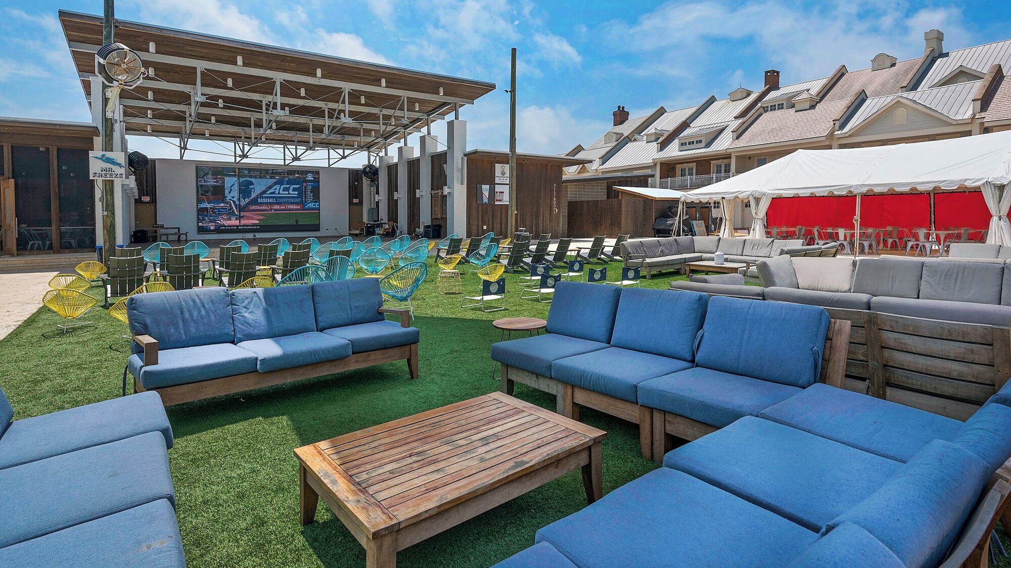 An outdoor seating area with blue sofas and chairs, a wooden table, and a covered area with a large screen displaying an event.