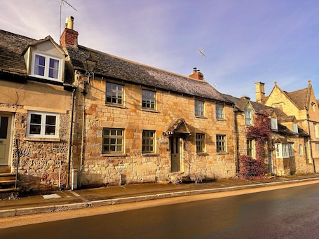 A traditional stone building with multiple windows and a chimney, located on a street with a sidewalk.