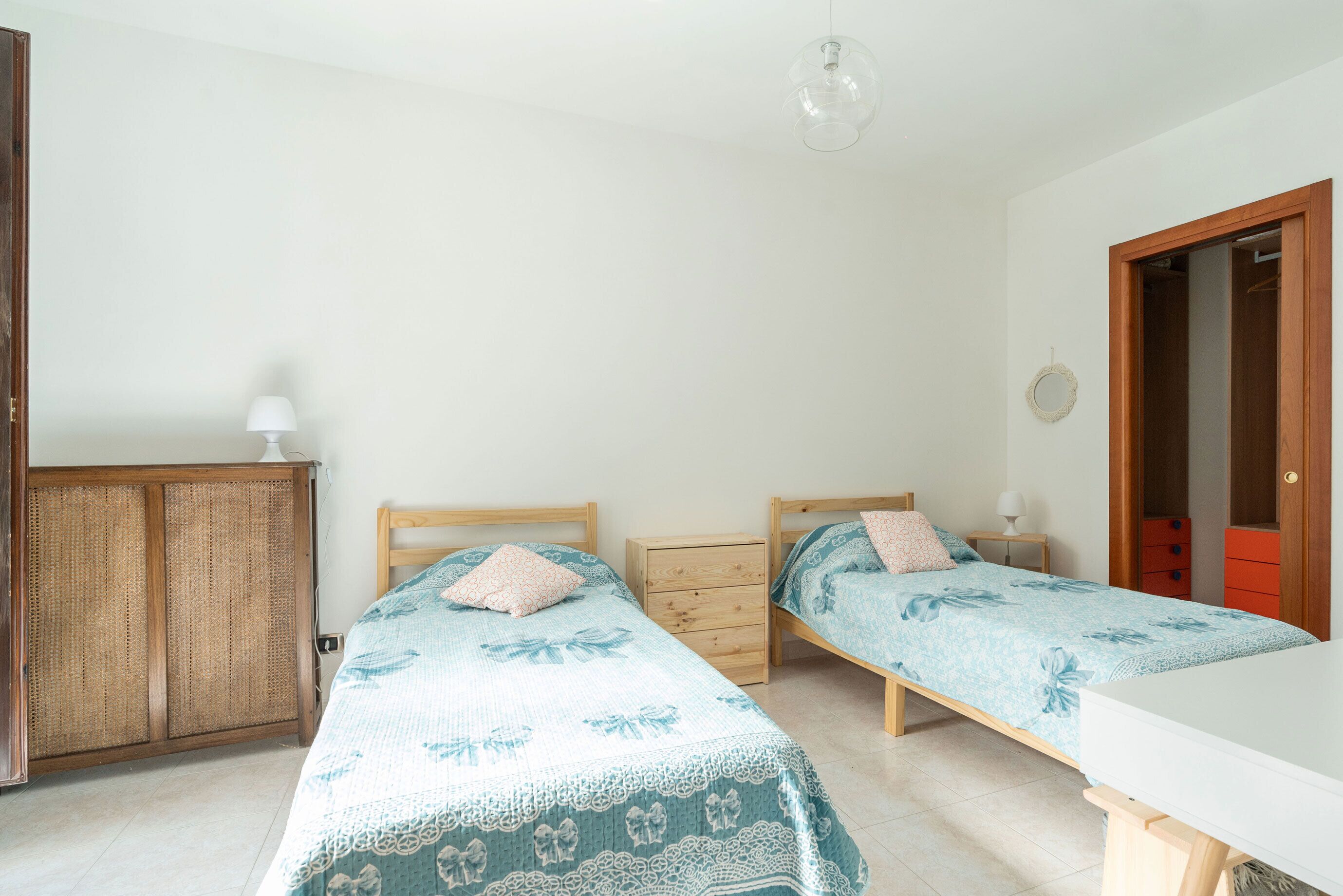 A bedroom with two single beds, a wooden wardrobe, a bedside table with a lamp, and a red cabinet.