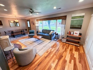A spacious living room with a wooden floor, a ceiling fan, a sofa, a chair, a coffee table, and a dining area with a bench. - Family Home Walk to Downtown | Near Zion & Bryce (Kanab)