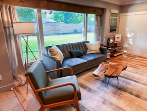A modern living room with a sofa, armchair, coffee table, and large windows overlooking a yard. - Family Home Walk to Downtown | Near Zion & Bryce (Kanab)