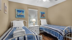 Een slaapkamer met twee bedden, elk voorzien van een blauw-wit gestreepte dekbedovertrek, witte hoofdborden en witte nachtkastjes. Twee aan de muur hangende schilderijen, waarvan één een zeilboot en de ander een vuurtoren voorstelt. In het midden is een raam met blinds te zien, en er hangt een plafondventilator.