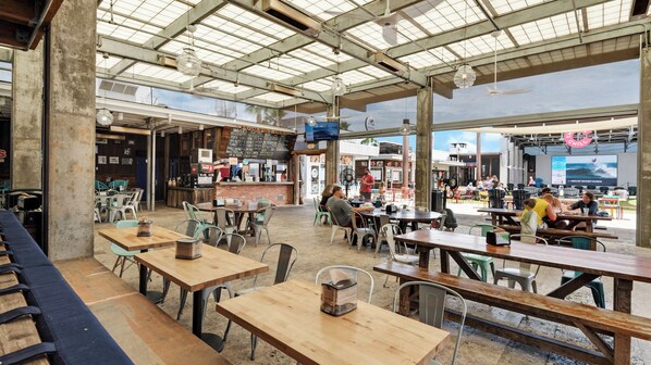 Un comedor interior con techos altos y luz natural, que incluye mesas y sillas de madera, una zona de bar y vistas al exterior.