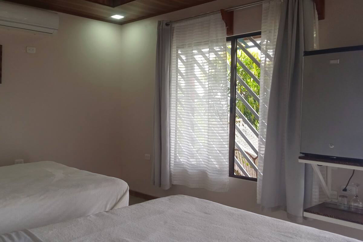 Une chambre avec deux lits simples, une fenêtre avec des rideaux légers, un climatiseur et une petite table avec une lampe.