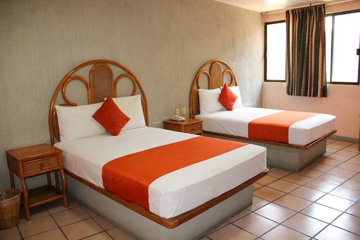 Zwei Einzelbetten mit Holz-Kopfteilen, weißer Bettwäsche und orangefarbenen Akzenten. Jedes Bett verfügt über einen Nachttisch und einen kleinen Korb.