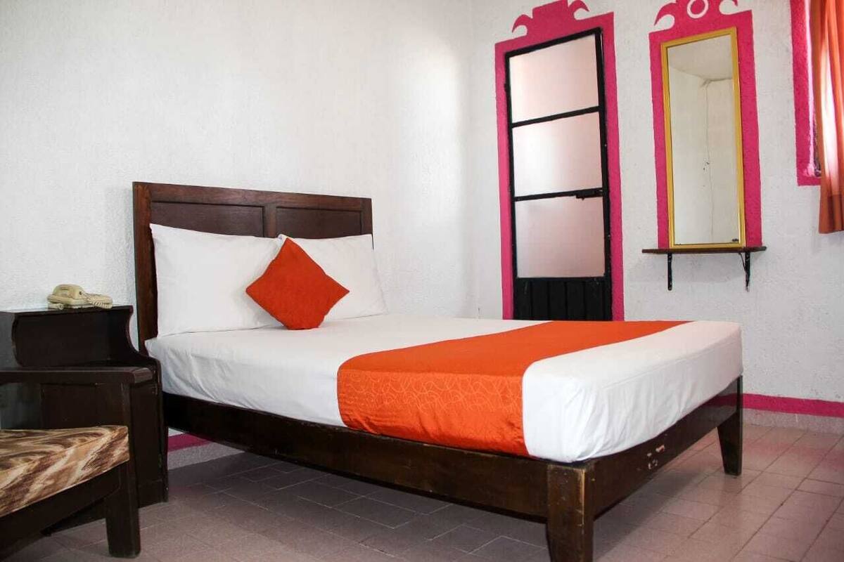 Ein Einzelbett mit Holzrahmen, weißer Bettwäsche und einer orangefarbenen Tagesdecke. Ein kleiner Nachttisch mit Lampe und Telefon ist sichtbar.