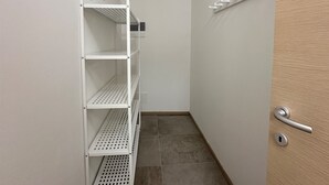 Un couloir étroit, avec une étagère métallique blanche adossée au mur gauche, une porte en bois sur la droite et un sol carrelé.