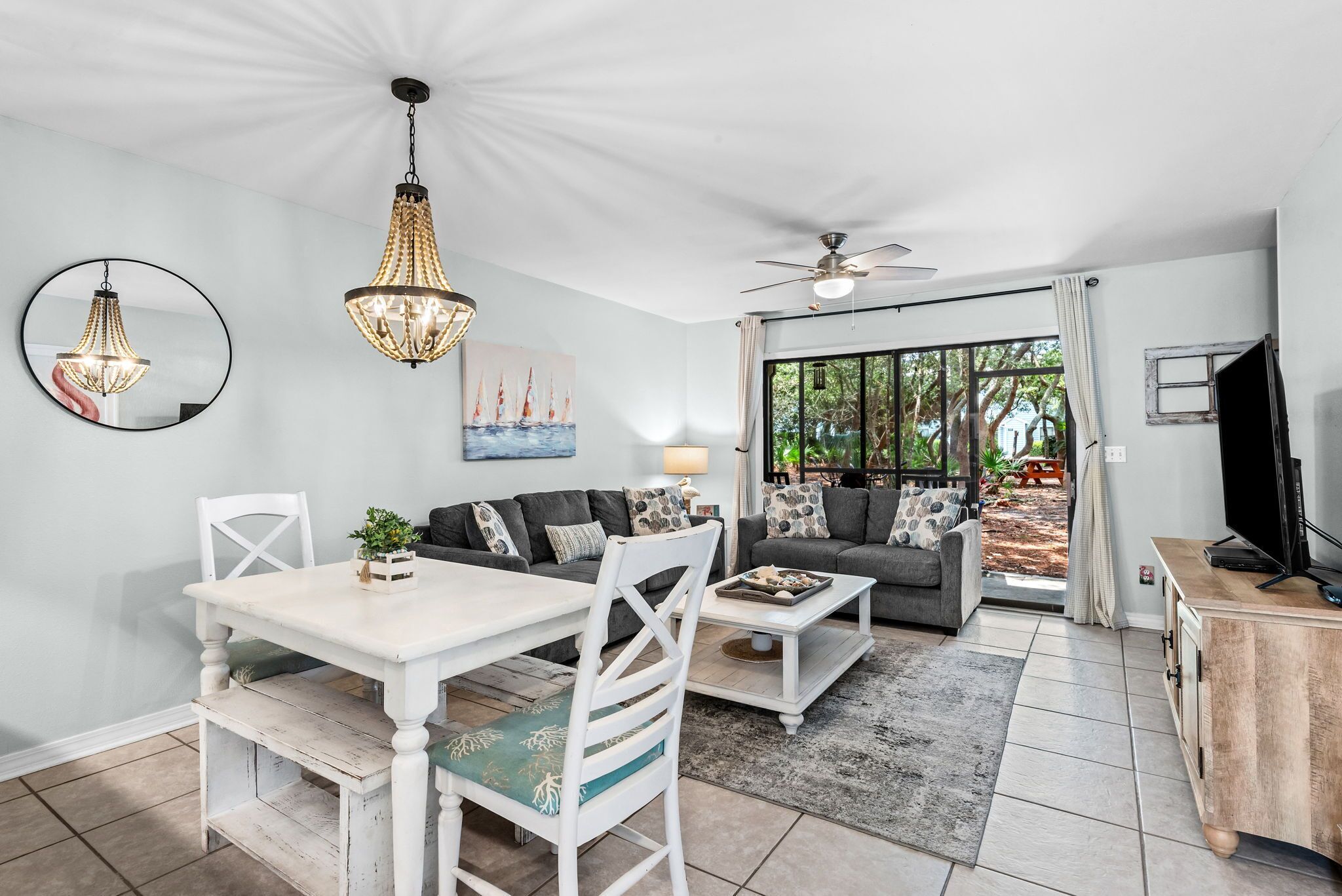 A modern living room with a dining area, a ceiling fan, and a large window with a view of greenery.