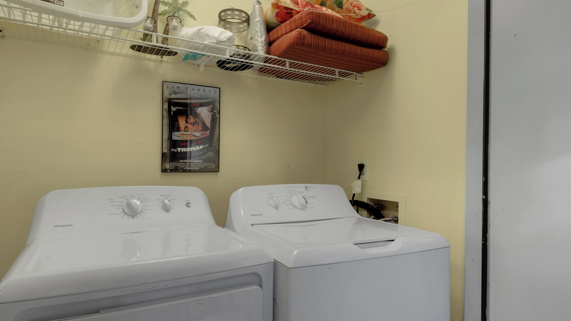 A laundry room with two front-loading washing machines, a poster on the wall, and a shelf above the machines.