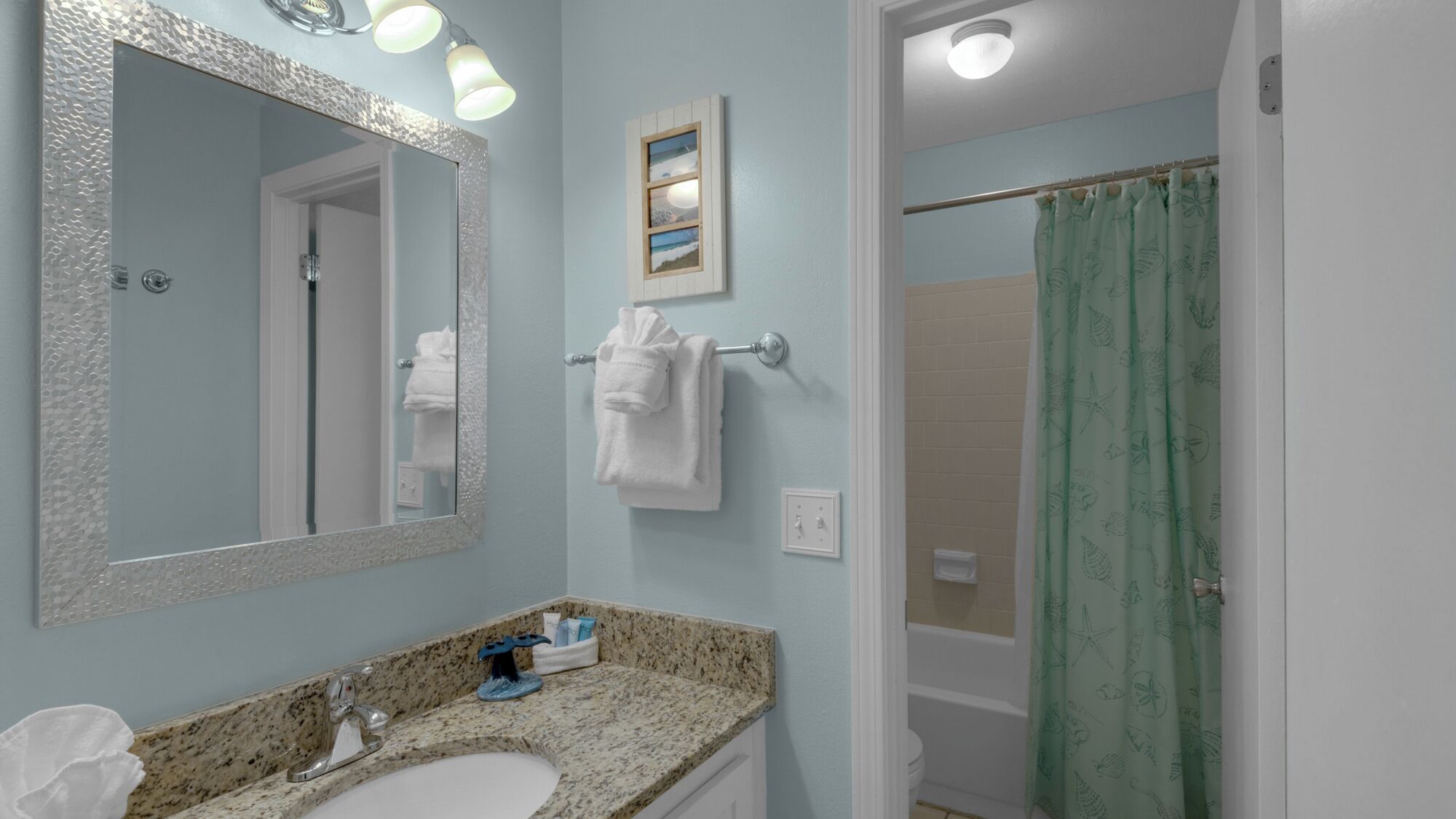 Un bagno con un piano di lavoro in granito, un lavabo bianco, uno specchio con illuminazione, un porta asciugamani e una tenda da doccia.
