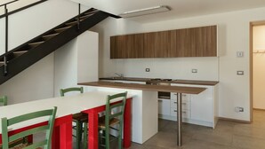 Een moderne keuken met een houten aanrecht, witte kasten en een ingebouwde kookplaat. Er is een eethoek met een witte tafel en groene stoelen. Op de achtergrond is een zwarte trap met houten treden te zien.