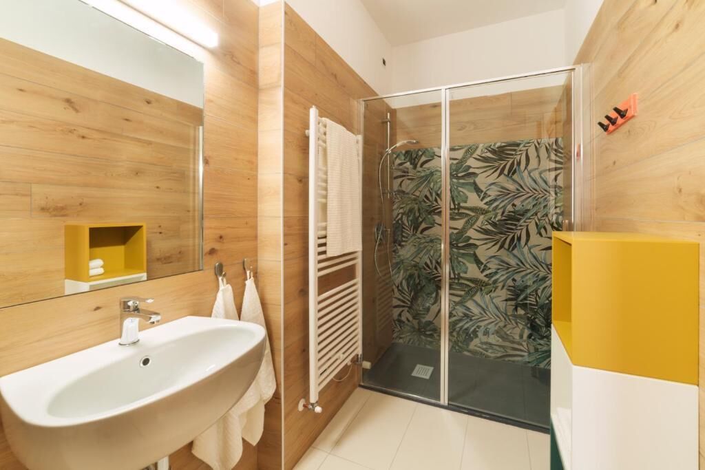 Un baño moderno con pared de madera, lavabo blanco, cabina de ducha con mampara de vidrio y un mueble amarillo.