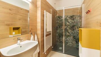 Un baño moderno con pared de madera, lavabo blanco, cabina de ducha con mampara de vidrio y un mueble amarillo.