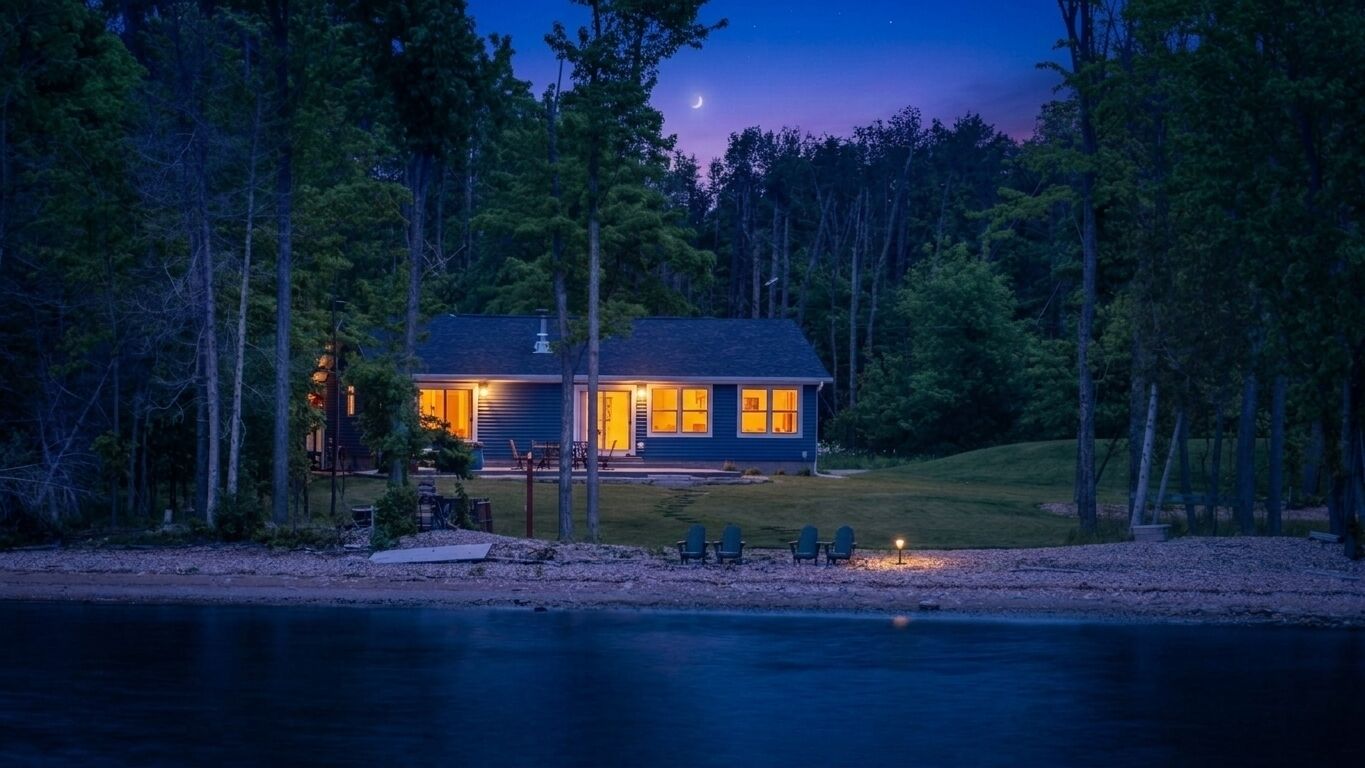 A cabin with a porch and outdoor seating, surrounded by trees and a body of water.
