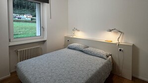 Un dormitorio con cama, dos lámparas de noche, una ventana con vistas a casas y vegetación, y un radiador.