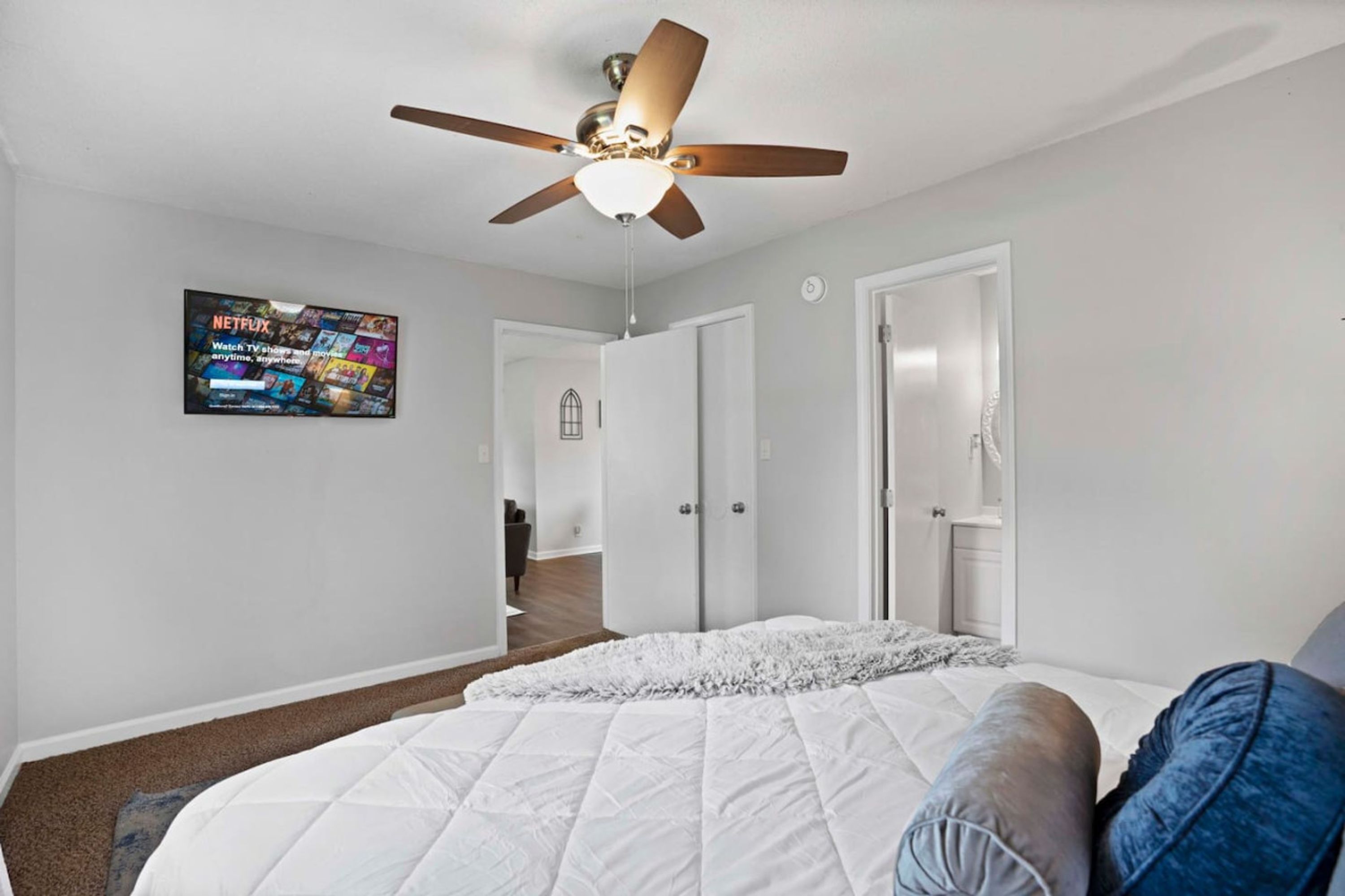 A bedroom with a bed, a ceiling fan, a wall-mounted TV, and a door leading to another room.