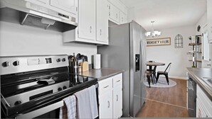 A modern kitchen with white cabinets, a stainless steel refrigerator, and a dining area with a table and chairs. - Charming House Close to Fort Campbell and Casino (Clarksville)