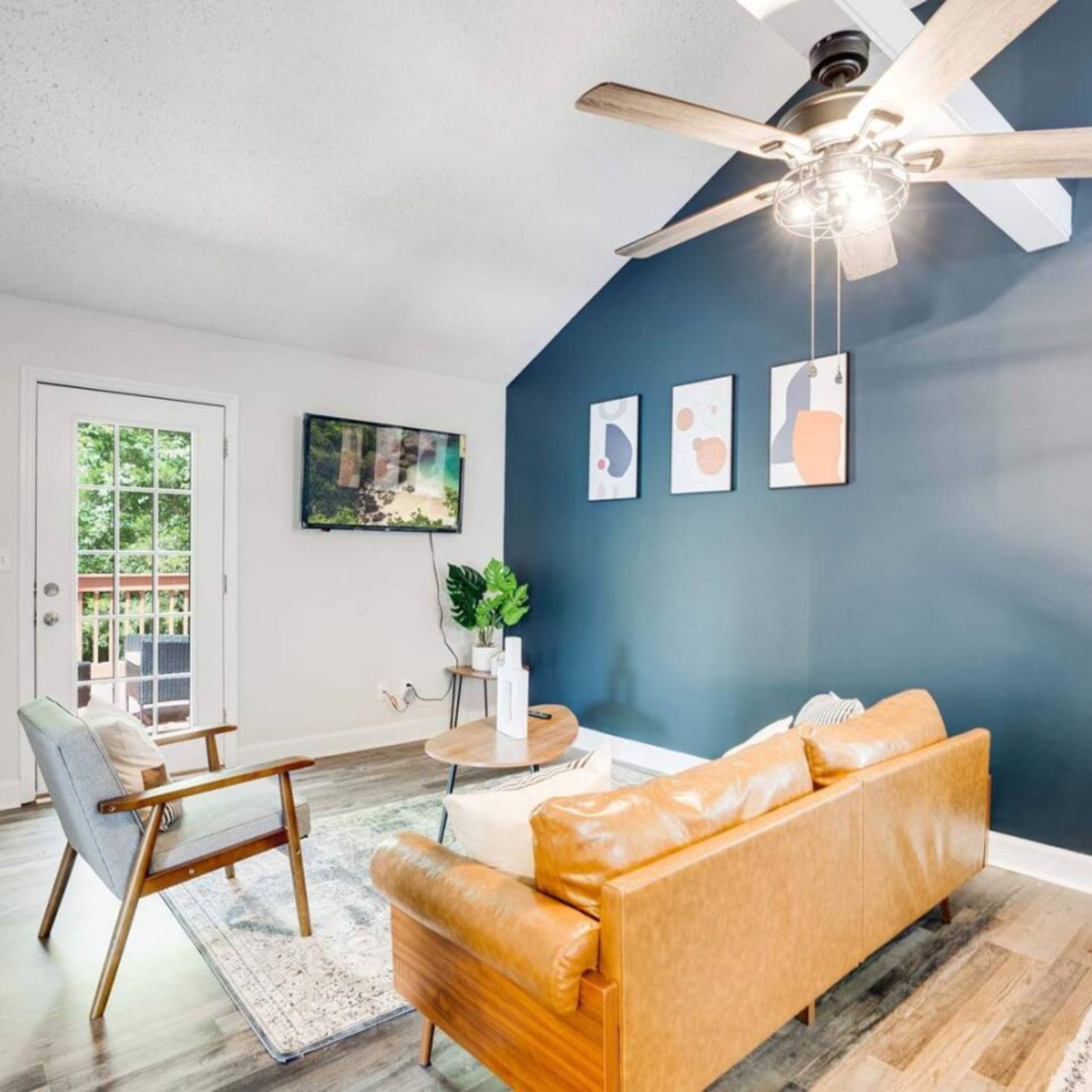 A modern living room with a blue accent wall, a ceiling fan, a sofa, a chair, a small table, and a TV mounted on the wall.