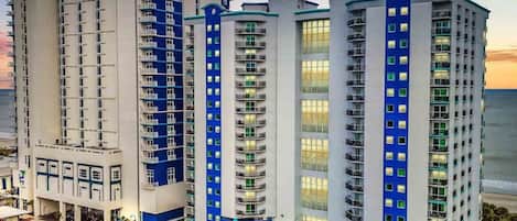 A multi-story hotel with a modern design, featuring balconies on each floor and a parking area in front.