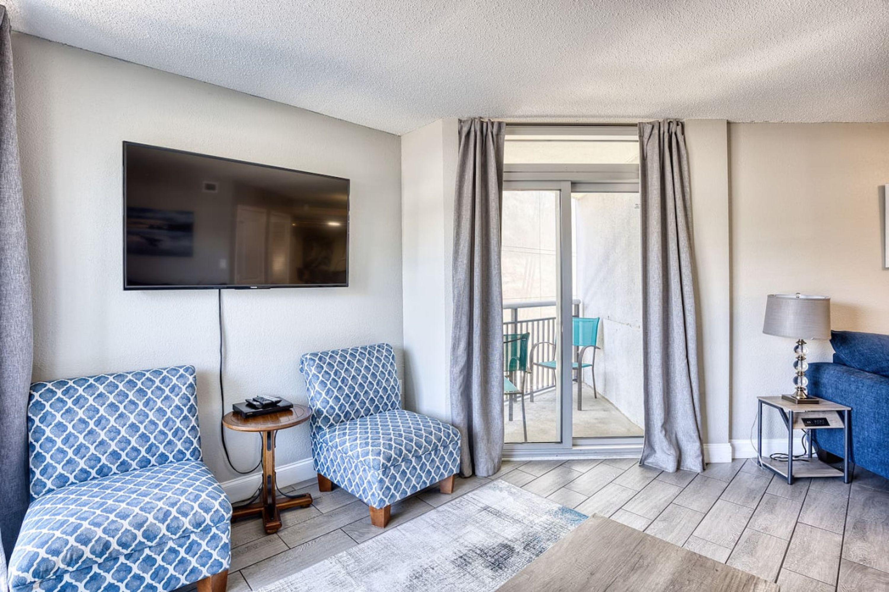 A modern hotel room with a large flat-screen TV mounted on the wall, a seating area with patterned blue chairs, a small round table, and a blue sofa.