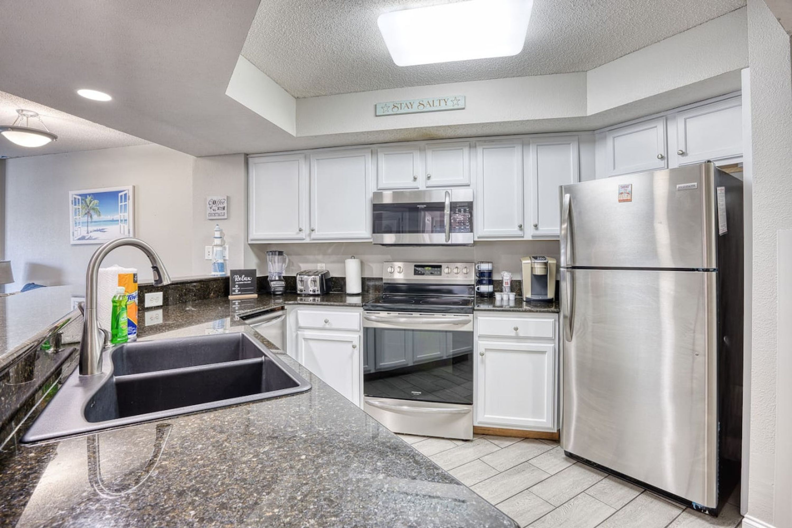 A modern kitchen with white cabinets, stainless steel appliances, a granite countertop, and a 'Stay Salty' sign.