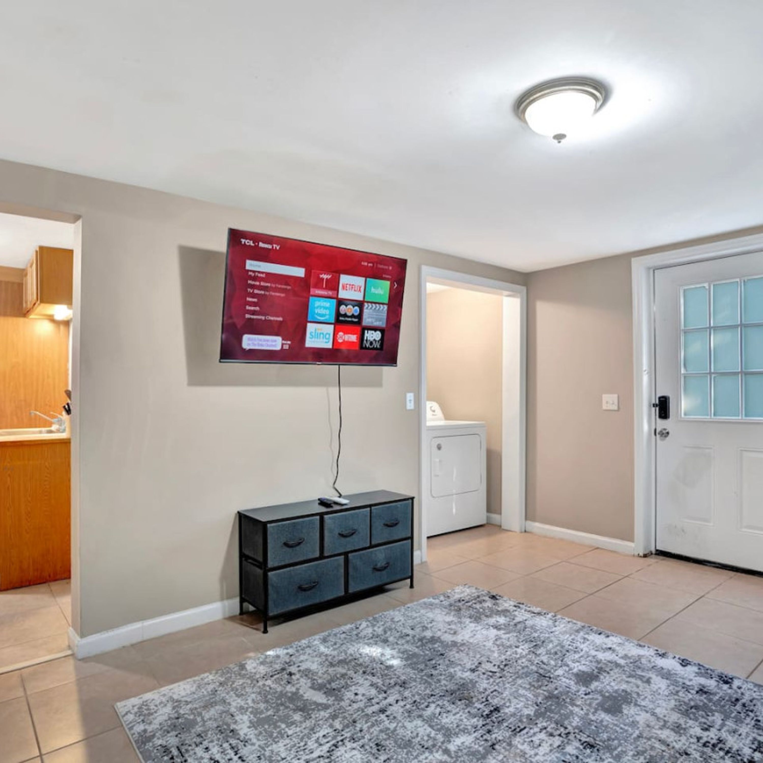 A living room with a flat-screen TV mounted on the wall, a dark-colored cabinet, a gray patterned rug, and a white door.