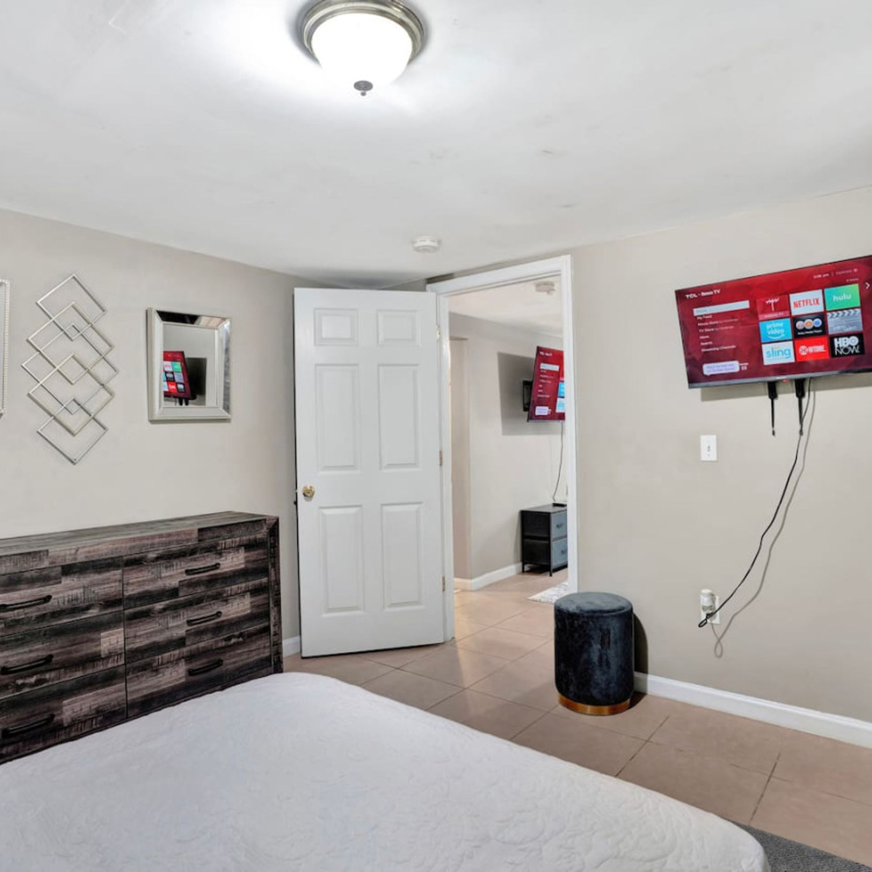 A bedroom with a bed, a dresser, a TV mounted on the wall, and a door leading to another room.