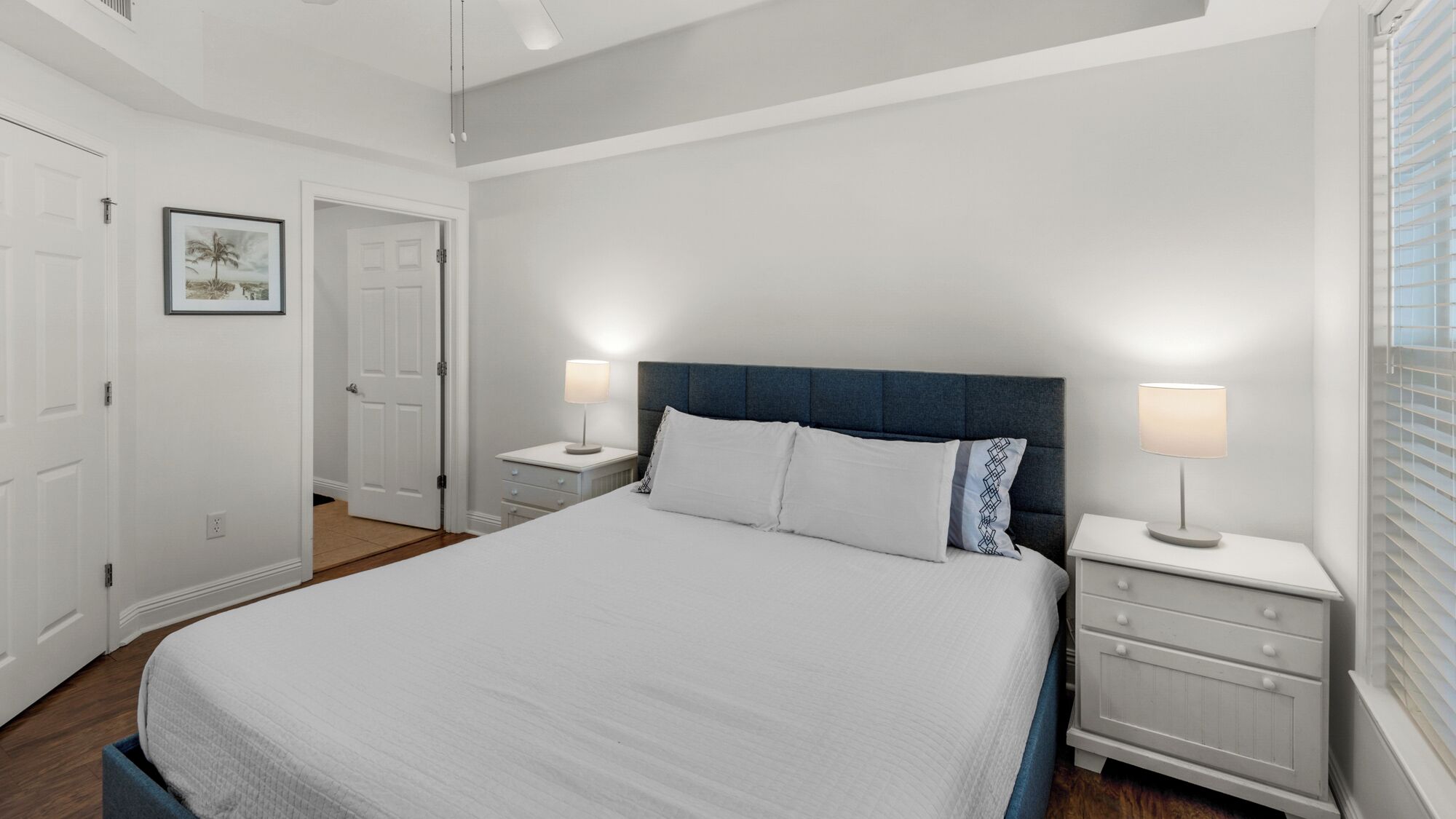 Un dormitorio con una cama grande, dos mesitas de noche con lámparas, una puerta y un cuadro enmarcado en la pared.
