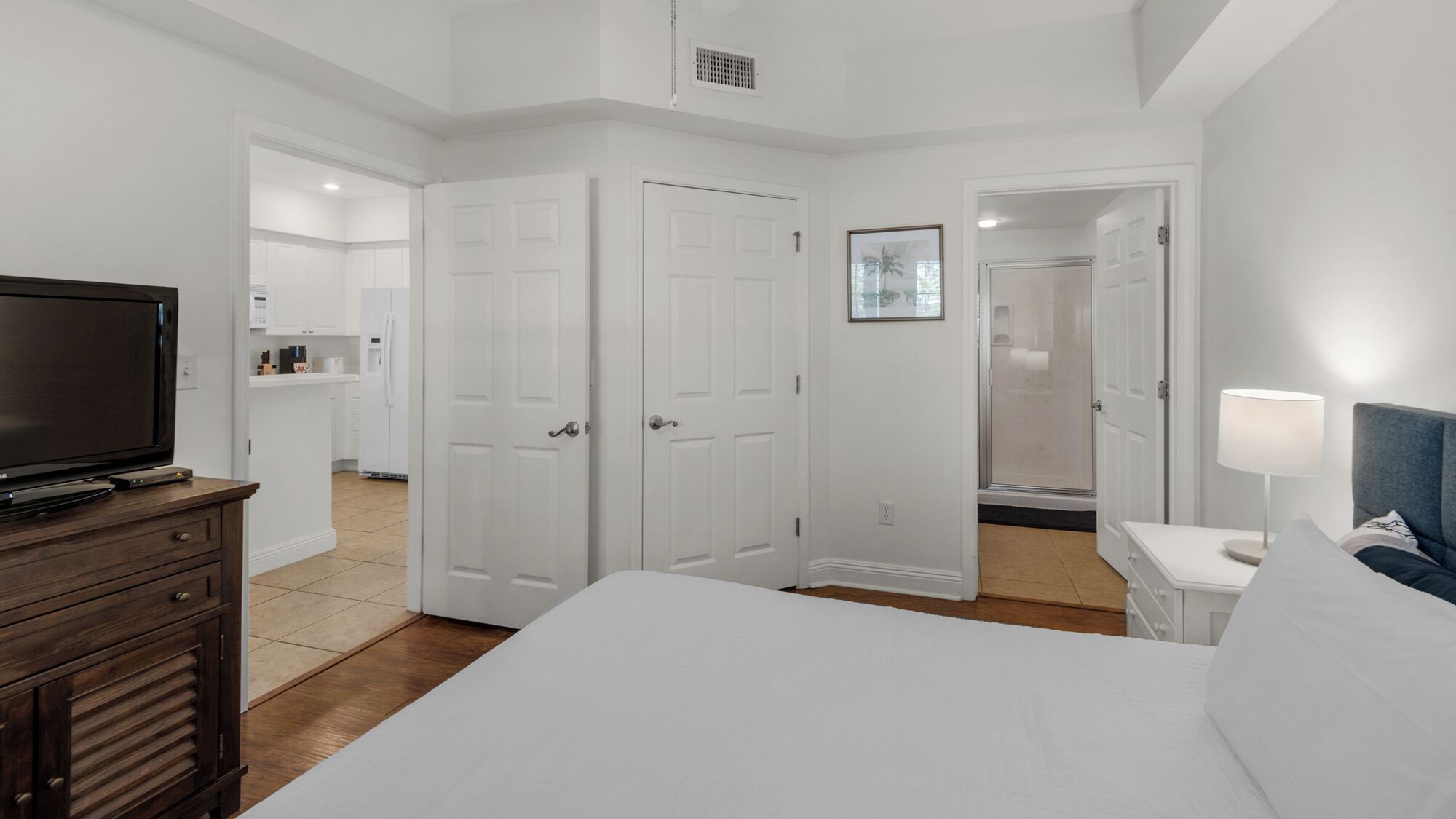 A hotel room with a large bed, a TV on a wooden dresser, a bathroom with a shower, and a nightstand with a lamp.