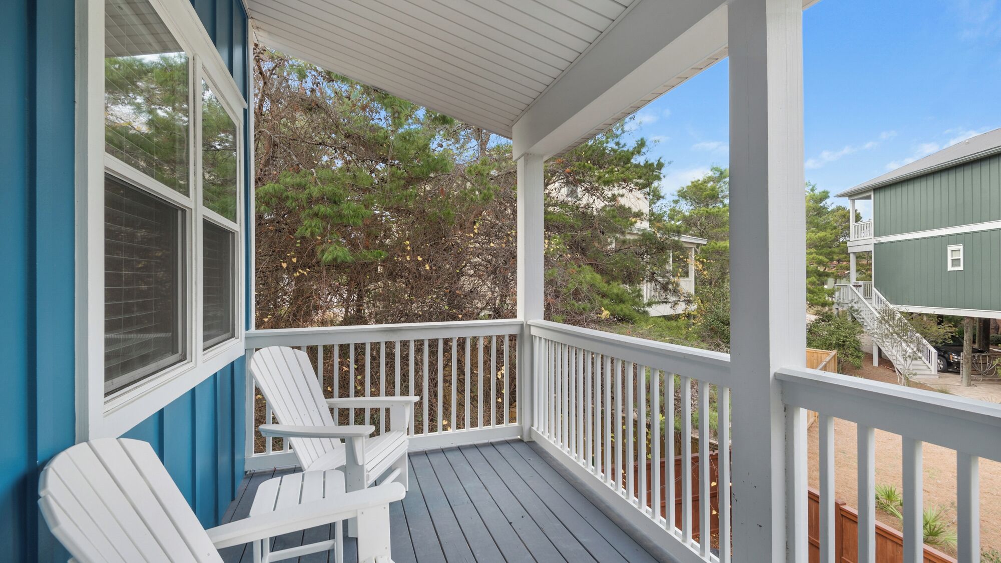 A covered porch with two white Adirondack chairs, a view of a green house, and a wooden deck.