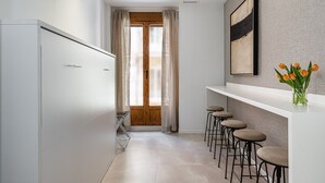 A narrow hallway with a white wardrobe, a wooden door, and a window with curtains. There are bar stools and a vase with flowers on a shelf.