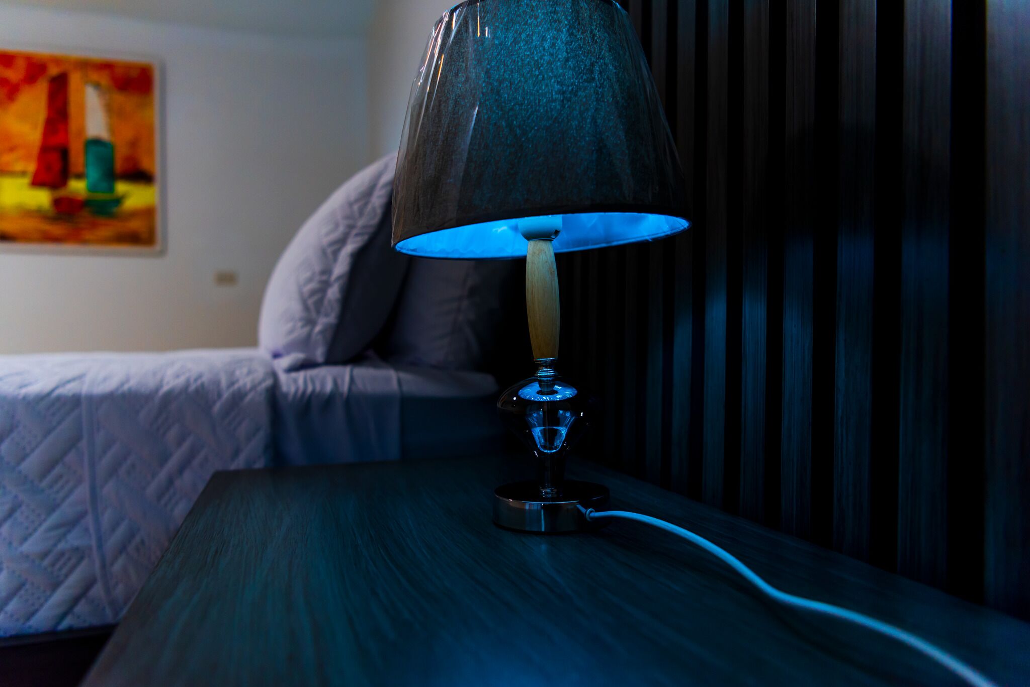 A bedside table with a lamp, a bed, and a painting on the wall.