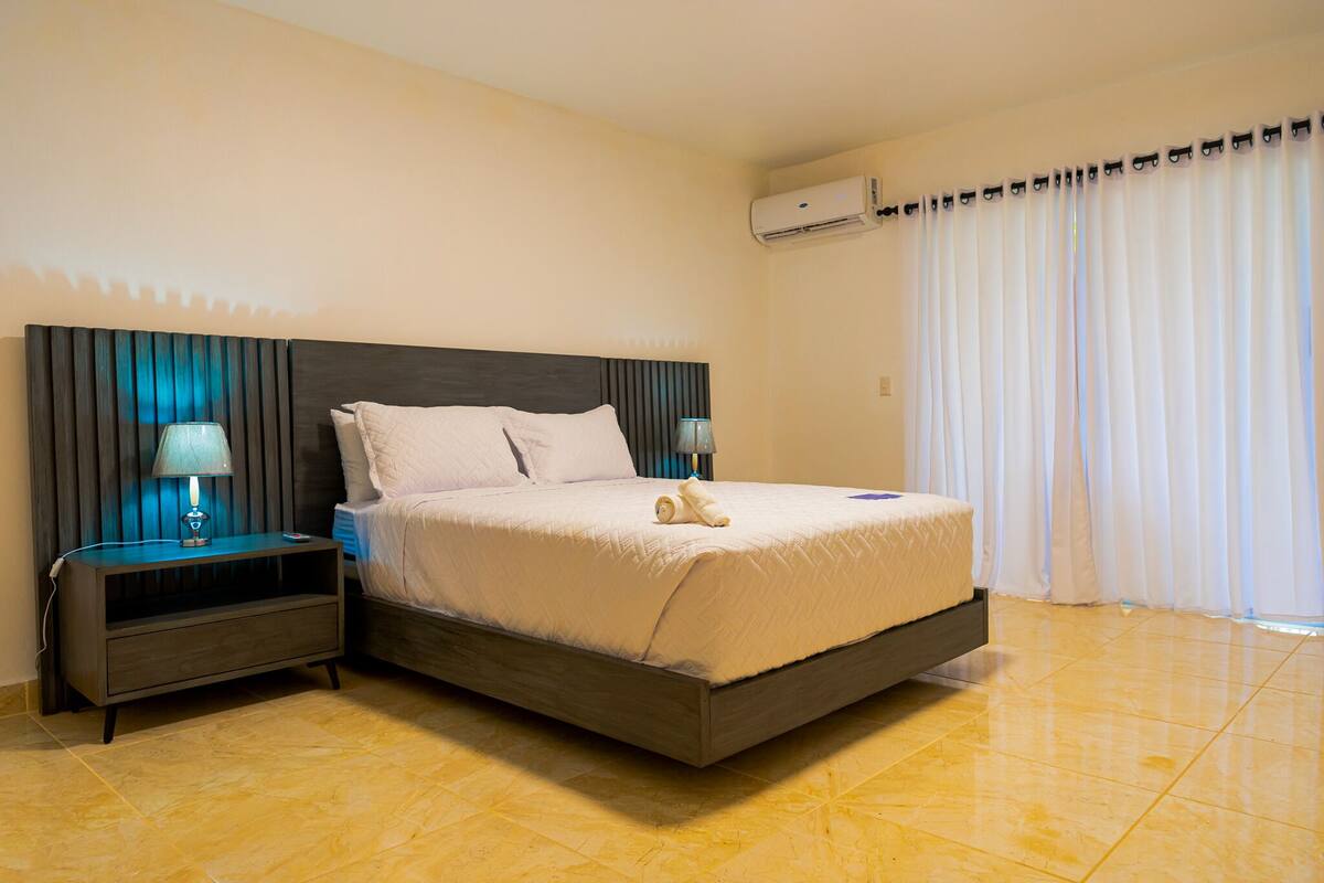 Una cama bien hecha con cabecera, una mesita de noche con lámpara y un equipo de aire acondicionado en la pared.