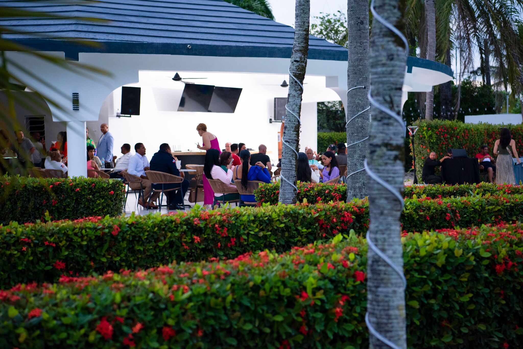 An outdoor gathering area with a white building, seating arrangements, and a well-maintained garden with hedges and flowers.