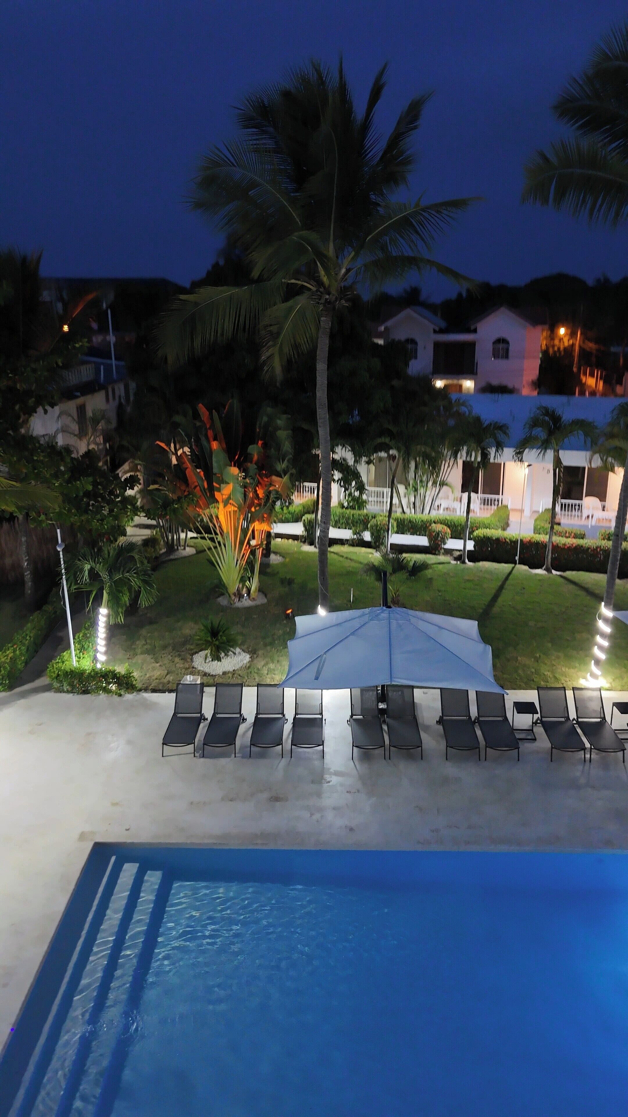 Un espace aménagé au bord de la piscine, avec des chaises longues et un grand parasol, entouré de palmiers et de pelouses bien entretenues.